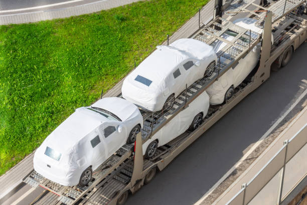 A car carrier truck transports several white, wrapped vehicles on a road next to a grassy area in MI, viewed from above.