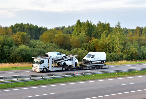 A car carrier truck from MI Auto Transport Lansing transports three white vans on a highway, with trees and greenery visible in the background under a cloudy sky.