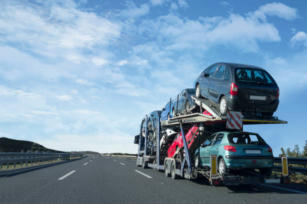 A car transporter truck carrying multiple cars drives on an empty highway under a bright blue sky with scattered clouds, showcasing the efficiency of Car Transport Lansing services in MI.