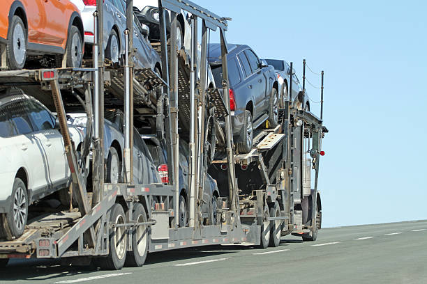 A MI Auto Transport Lansing car carrier truck loaded with multiple vehicles drives up a road under a clear blue sky.