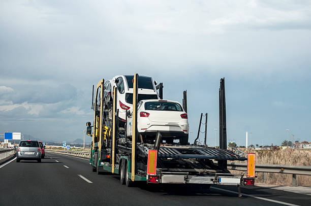 A car carrier truck transports several white vehicles along a multi-lane highway in Lansing, MI, with other cars visible on the road and distant buildings under a cloudy sky—capturing the essence of MI Car Shipping Lansing.